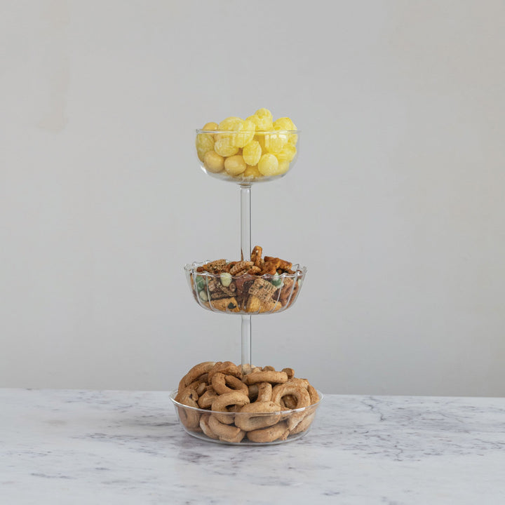 Three-tiered glass snack stand with snacks on a marble surface and gray background