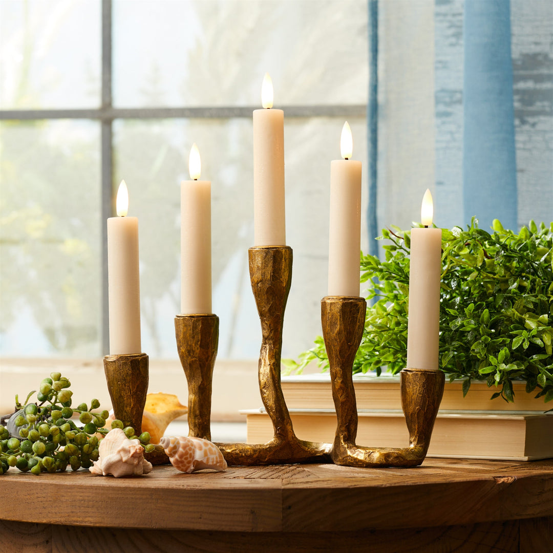 Set of five lit candles in decorative brass candle holders on a wooden surface with a blurred window and plant in the background.