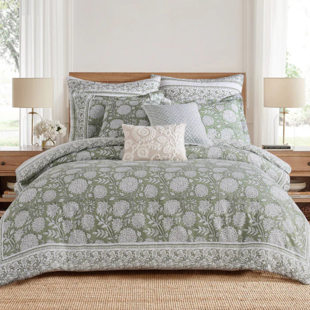 A neatly made bed in a bright bedroom setting, featuring a sage green and white floral patterned comforter set with matching pillows, a textured cream accent pillow, and a wooden headboard.