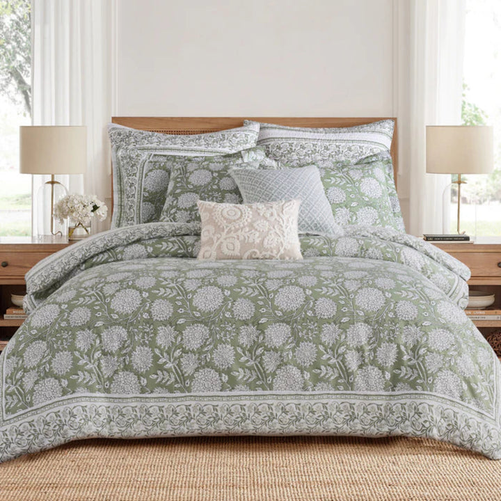 A neatly made bed in a bright bedroom setting, featuring a sage green and white floral patterned comforter set with matching pillows, a textured cream accent pillow, and a wooden headboard.