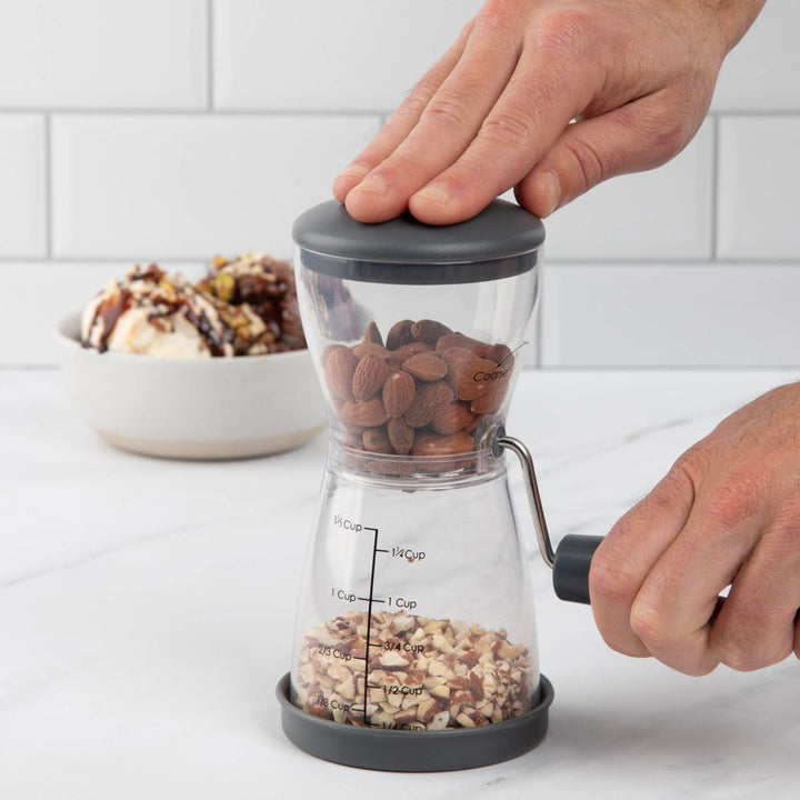 A clear manual nut cracker with a dark gray lid and handle, being operated by two hands. Almonds and chopped nuts are visible inside the transparent chamber, set on a white marble countertop with a white subway tile backsplash and a bowl of dessert in the background.