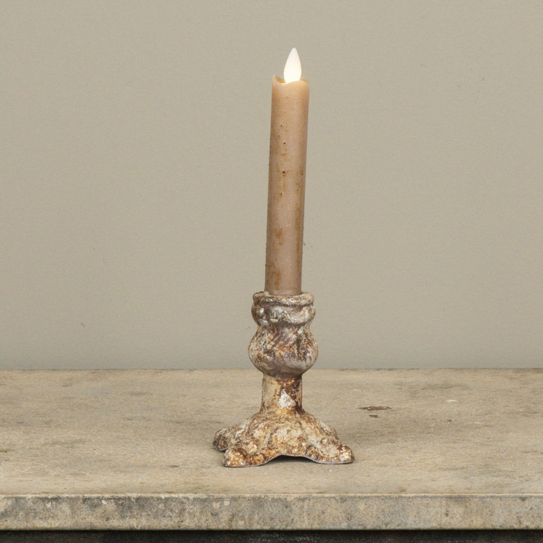 A single lit taper candle in a rustic, textured metal holder with a weathered silver and gold finish, set against a neutral beige background.