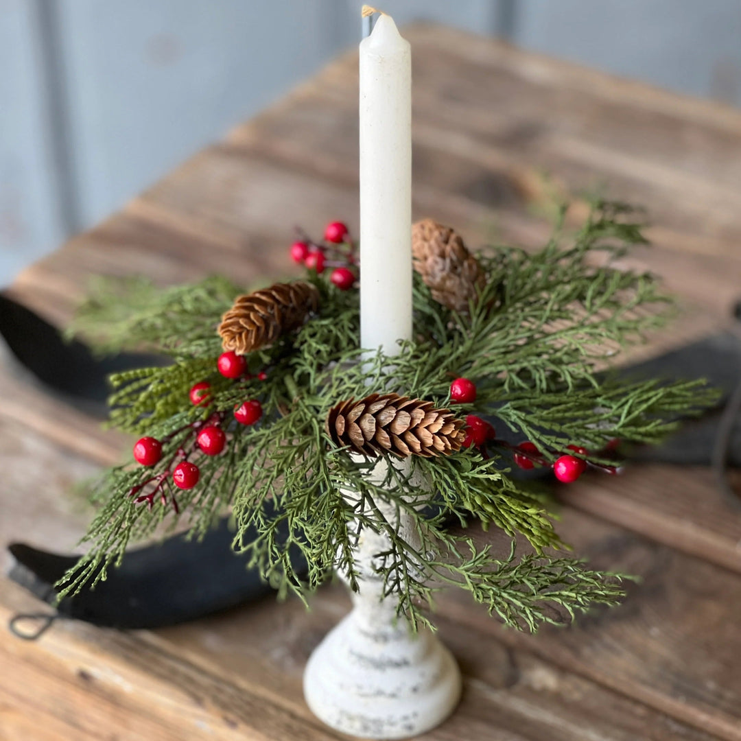 Decorative candle holder with greenery, pine cones, and red berries on a wooden surface.