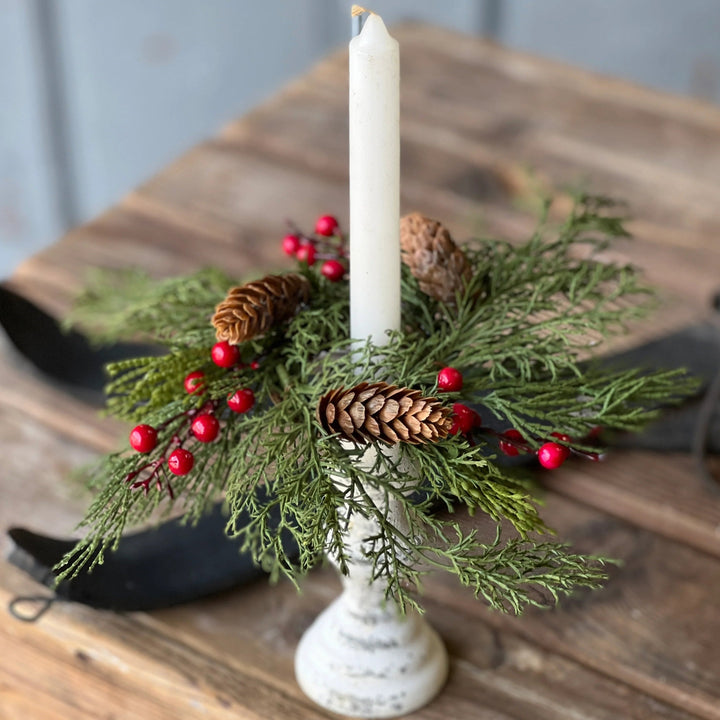 Decorative candle holder with greenery, pine cones, and red berries on a wooden surface.