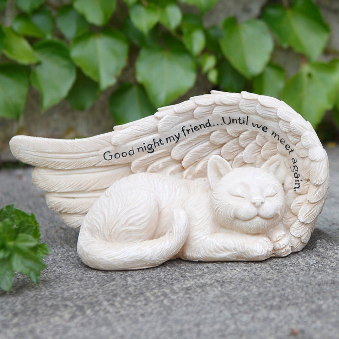 White cat figurine with angel wings on a stone surface with green foliage in the background