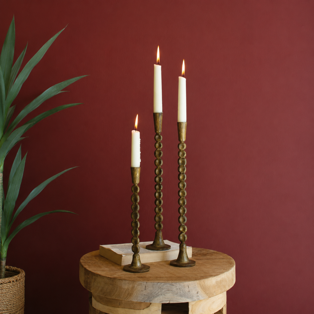 Three brass spiral candlestick holders with lit white taper candles, styled on a round wooden side table against a dark red wall with a potted plant on the left.