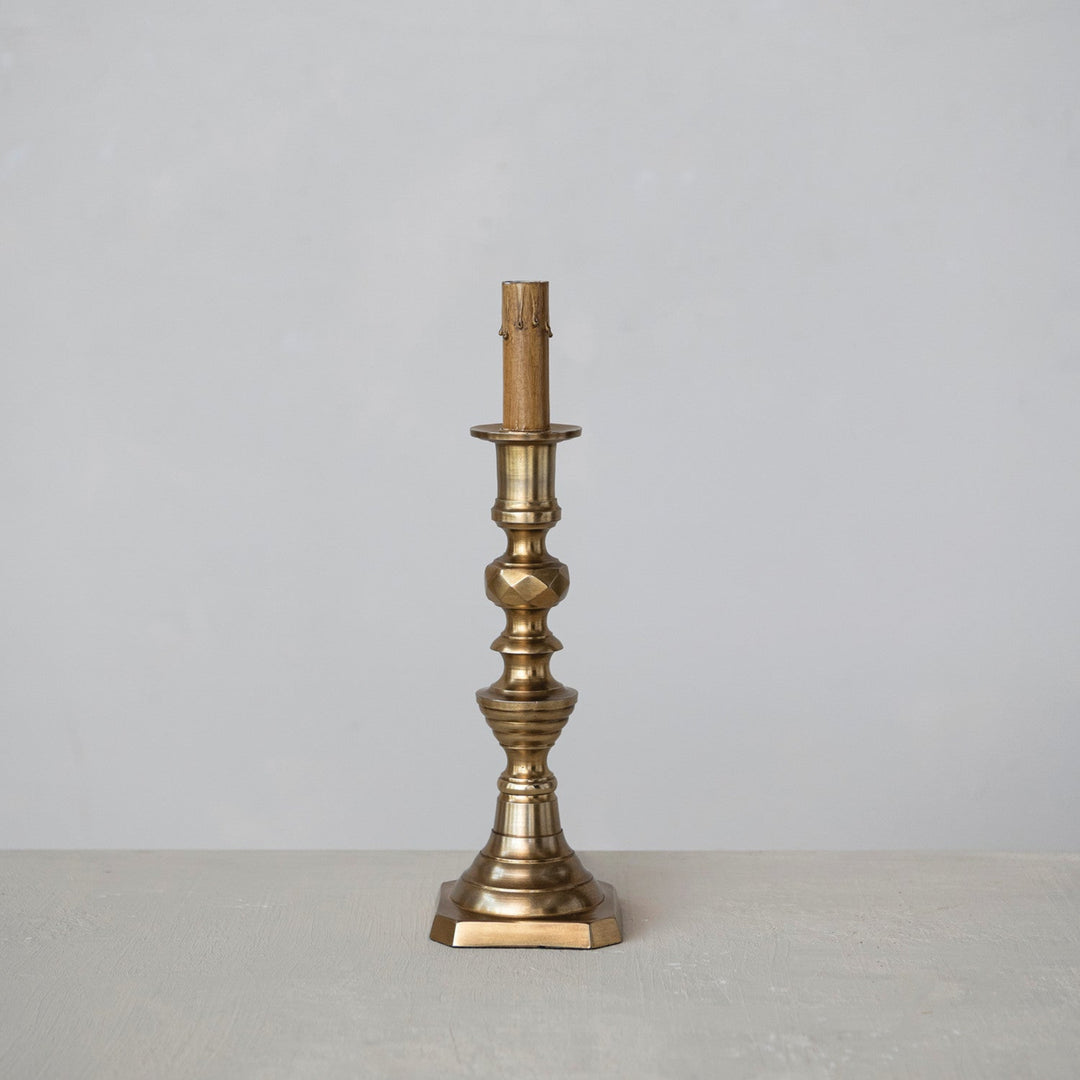 A single brass candlestick holder with an ornate fluted base and a candle sleeve, standing on a light gray surface against a neutral gray background.