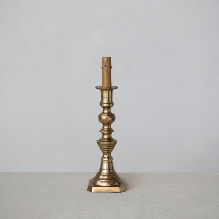 A single brass candlestick holder with an ornate fluted base and a candle sleeve, standing on a light gray surface against a neutral gray background.