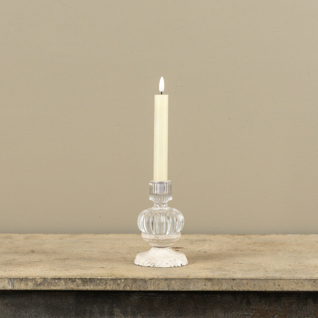 Clear glass candle holder with a lit candle on a wooden surface against a beige wall.