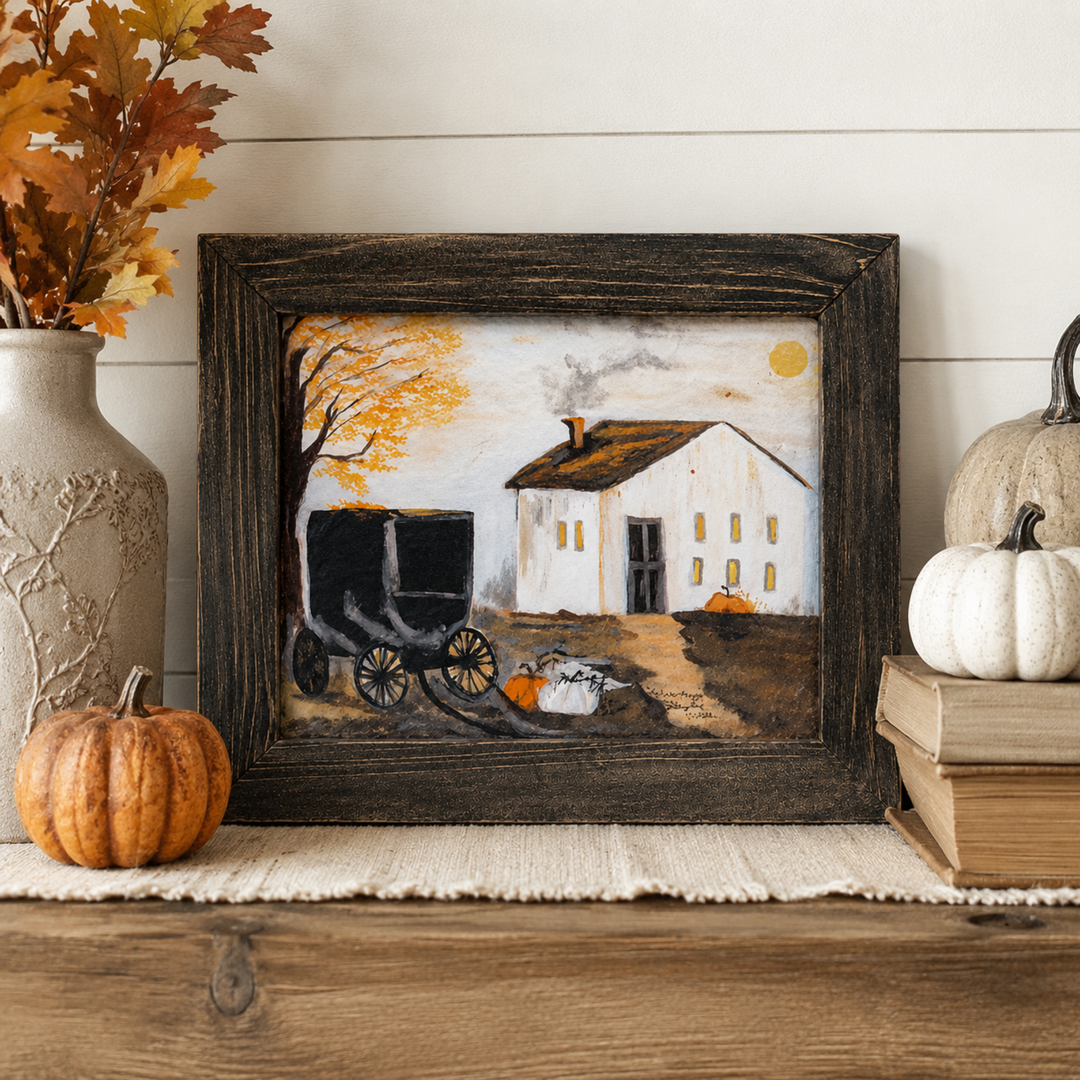 A framed autumn-themed art print featuring a white farmhouse, a black carriage, and an orange tree, styled on a wooden shelf with pumpkins, books, and a vase.