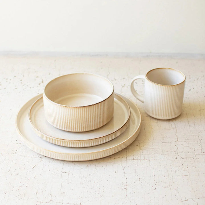 A set of cream-colored ceramic tableware, including a stack of plates and a ribbed bowl on the left, and a ribbed mug on the right, photographed on a textured light beige surface against a neutral off-white background.