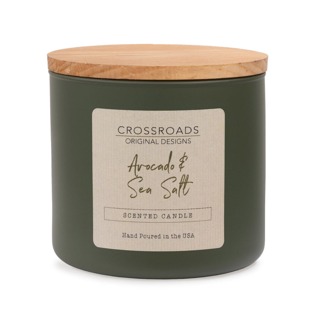 A dark olive green scented candle jar with a light natural wood lid and a cream-colored paper label. The label reads 'CROSSROADS ORIGINAL DESIGNS' and 'Avocado & Sea Salt'.