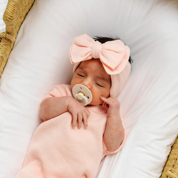 Newborn baby in a pink outfit with a large bow headband and pacifier, lying on a white blanket.