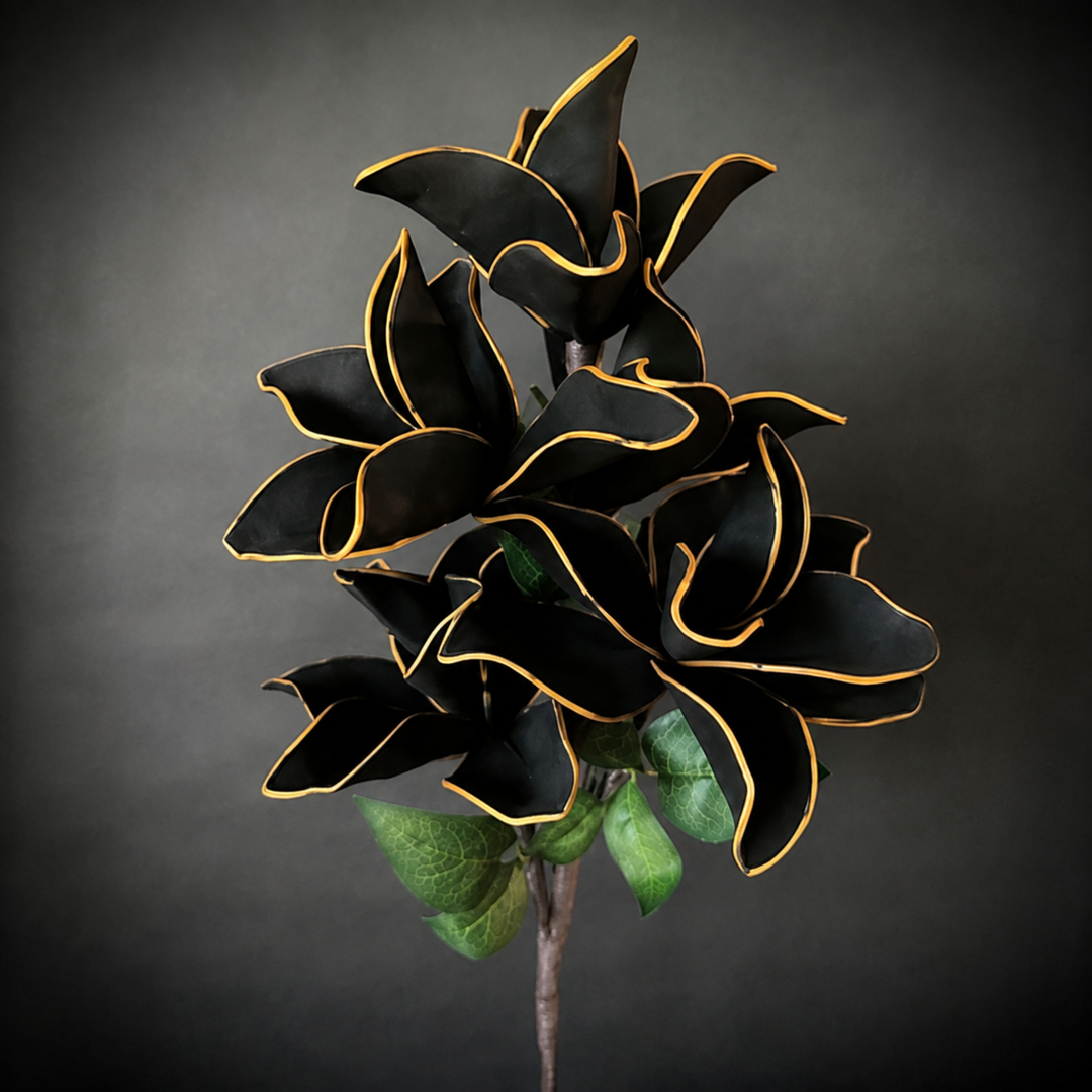 A decorative artificial flower stem featuring large black petals with thin gold edging and a few green leaves, photographed against a dark gray studio background.
