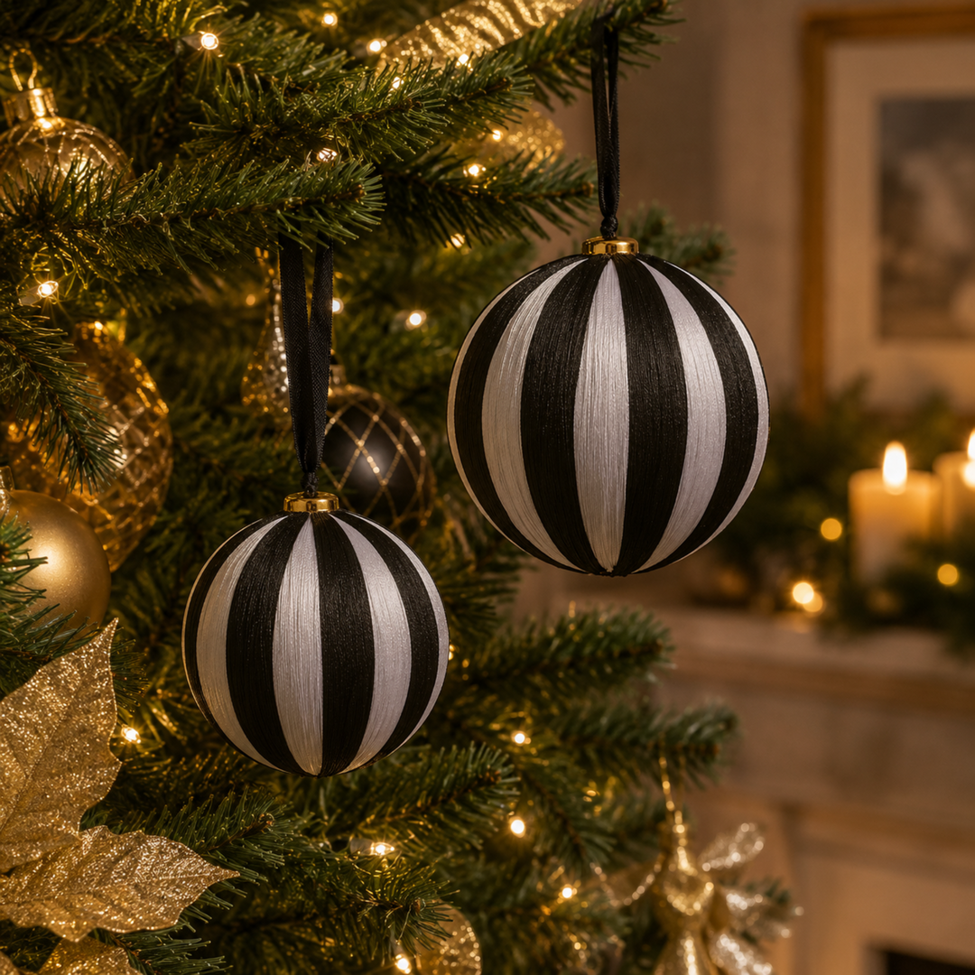 Two black and white striped spherical Christmas ornaments hanging on a decorated green Christmas tree with warm lights and gold ornaments in the background.