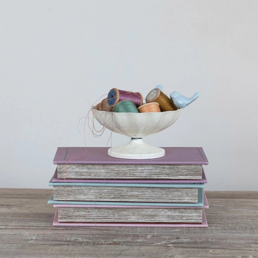 Stack of books with a decorative bowl containing colorful spools of thread on a wooden surface.