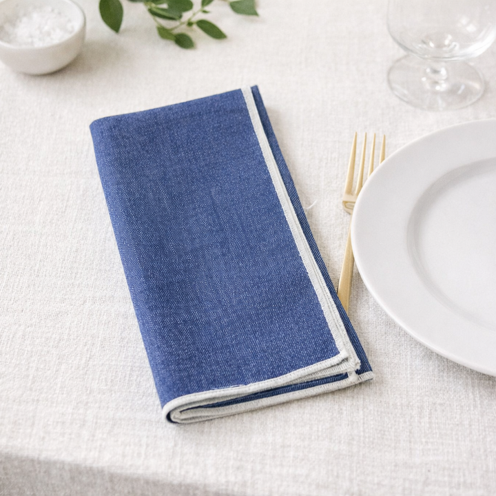 Folded blue napkin on a white tablecloth with a plate, fork, and knife.