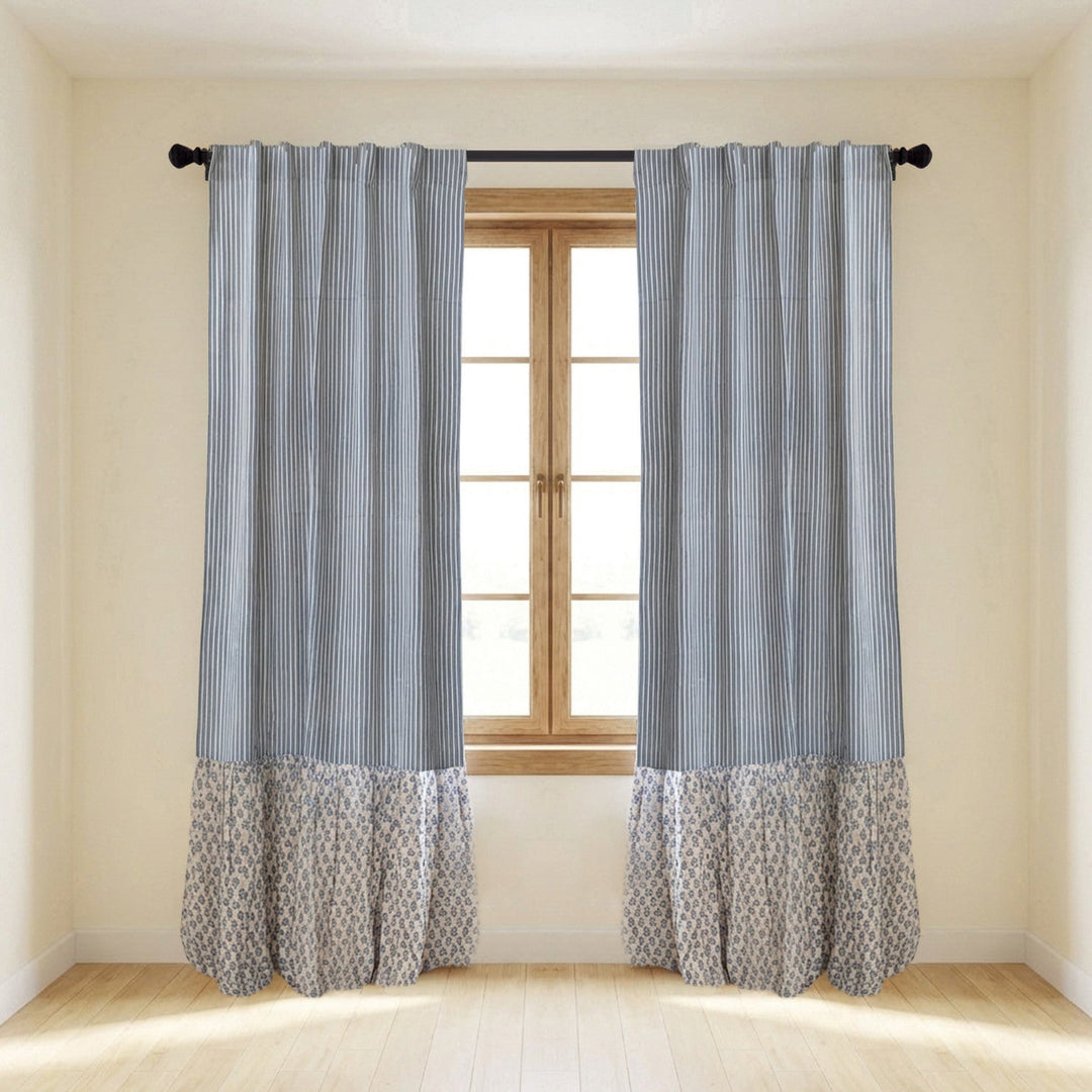 A pair of gray and white striped curtain panels with a patterned ruffled hem, hanging on a black rod over a window in a bright room.