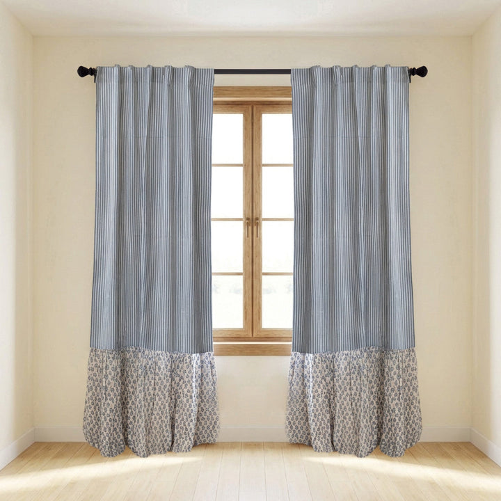 A pair of gray and white striped curtain panels with a patterned ruffled hem, hanging on a black rod over a window in a bright room.