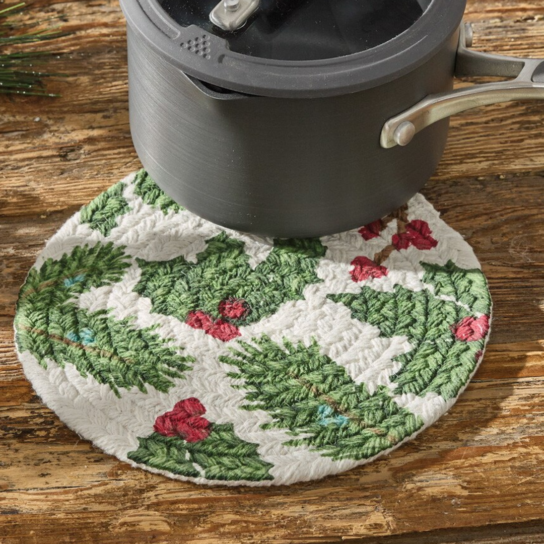 A round quilted white pot holder with a green holly and red berry pattern, shown under a dark gray pot on a rustic wooden table.