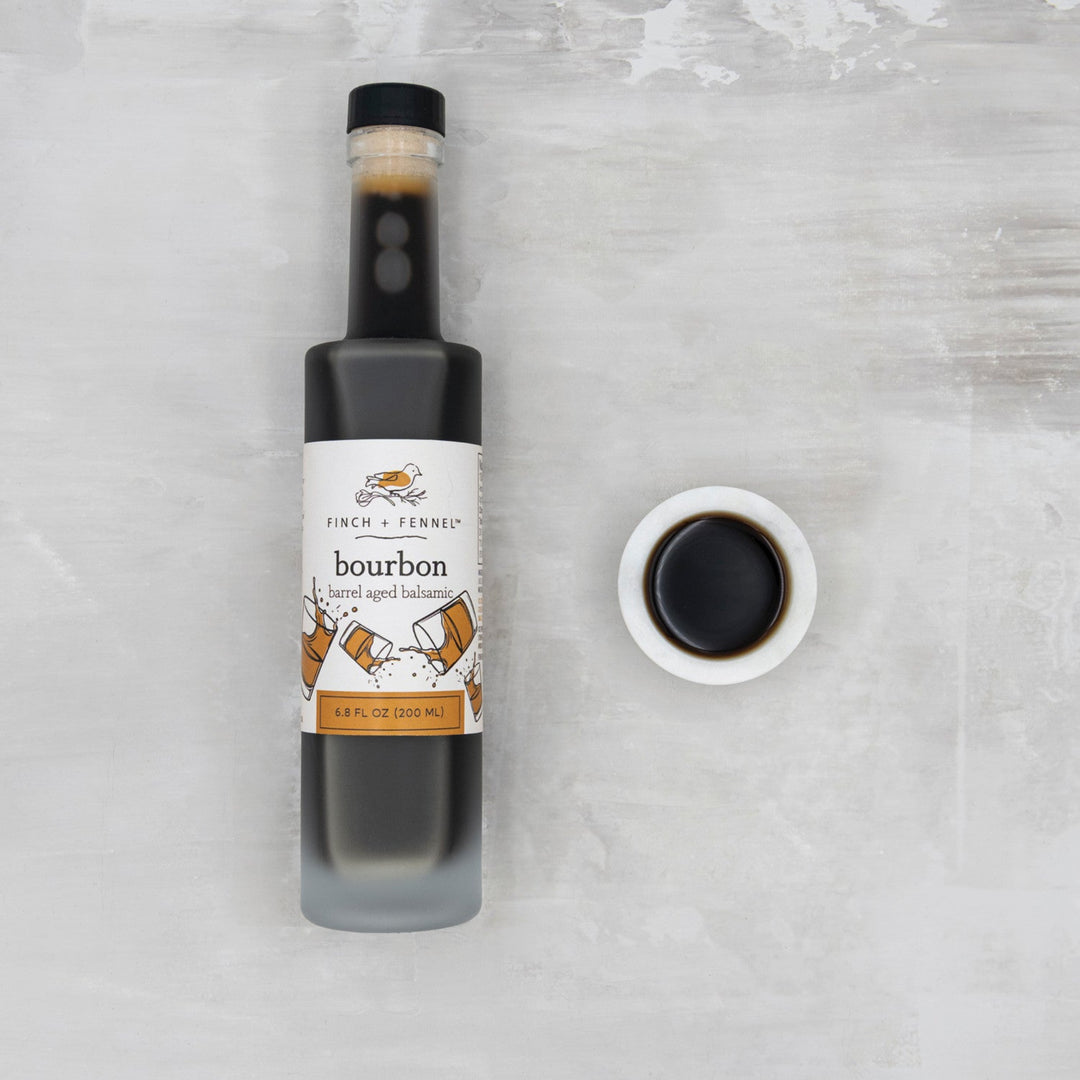 Bottle of bourbon balsamic vinegar with a small bowl of the product on a textured gray background