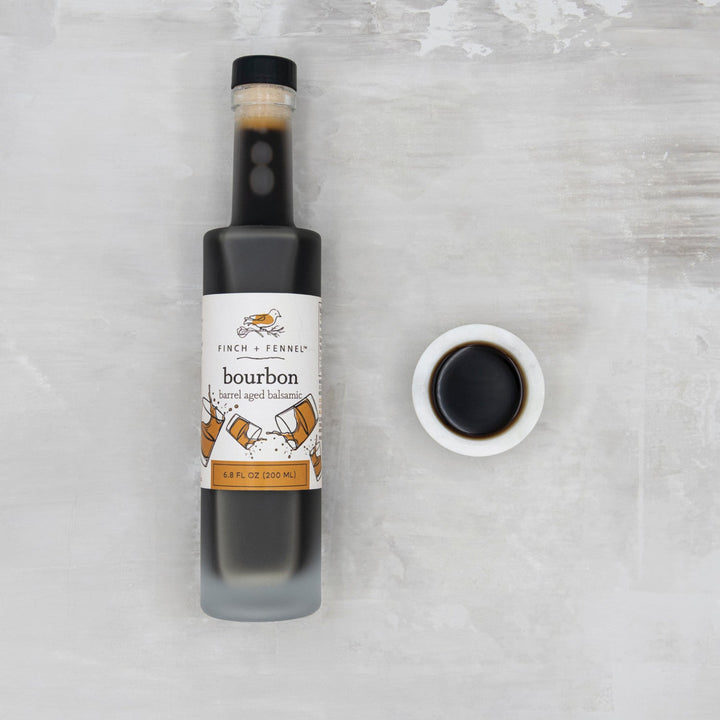 Bottle of bourbon balsamic vinegar with a small bowl of the product on a textured gray background