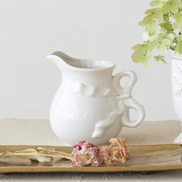 A white ceramic pitcher with a decorative embossed bow on the front and a loop handle on the side, styled on a wooden tray with dried flowers and a vase of green leaves in the background.