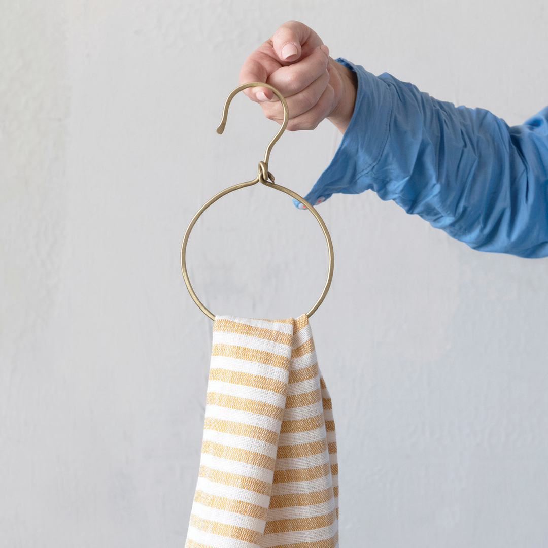 A hand wearing a blue long-sleeve shirt holds a brass ring hanger with a beige and white striped cloth hanging from it against a light gray wall.