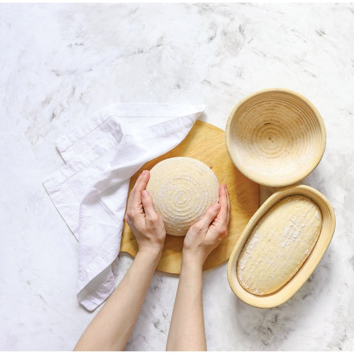 Hands handling a round loaf of bread on a wooden board with two other loaves in woven baskets on a marble surface.