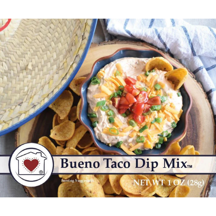 Bueno Taco Dip Mix in a blue bowl filled with creamy dip, shredded cheese, diced tomatoes, and green onions, surrounded by tortilla chips on a wooden board with a straw hat and striped cloth in the background.
