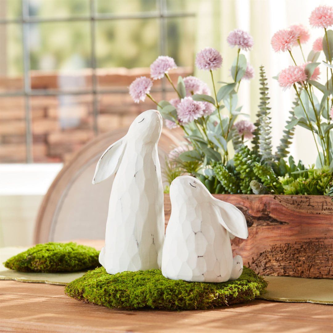 Two white marble rabbit sculptures on a bed of green moss with pink flowers in the background.