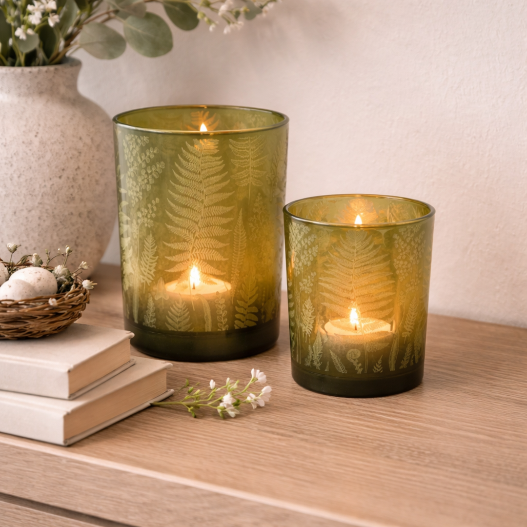 Two green glass candle holders with lit candles on a wooden surface, accompanied by a vase, books, and flowers.