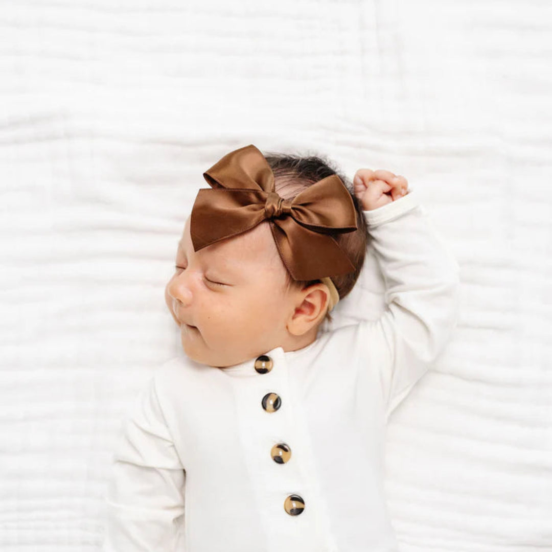A sleeping newborn baby wearing a white long-sleeve bodysuit with gold buttons and a brown satin bow headband, lying on a white textured blanket.
