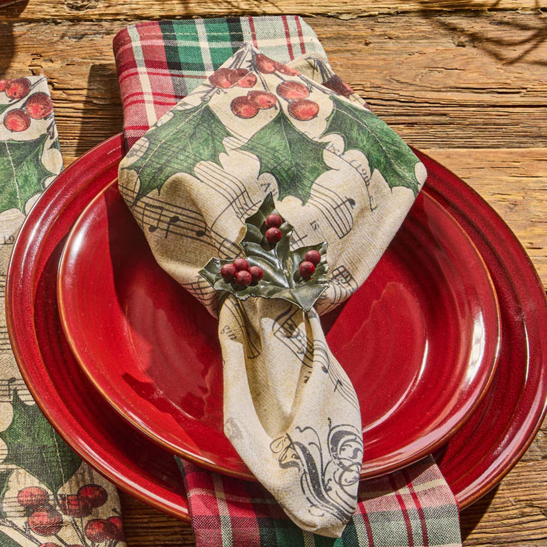 A styled holiday table setting featuring a cream napkin printed with green holly leaves, red berries, and black musical notes, tied with a silver holly pin. The napkin is layered over a stack of red ceramic plates on a rustic wooden table, with a matching plaid napkin partially visible on the left.