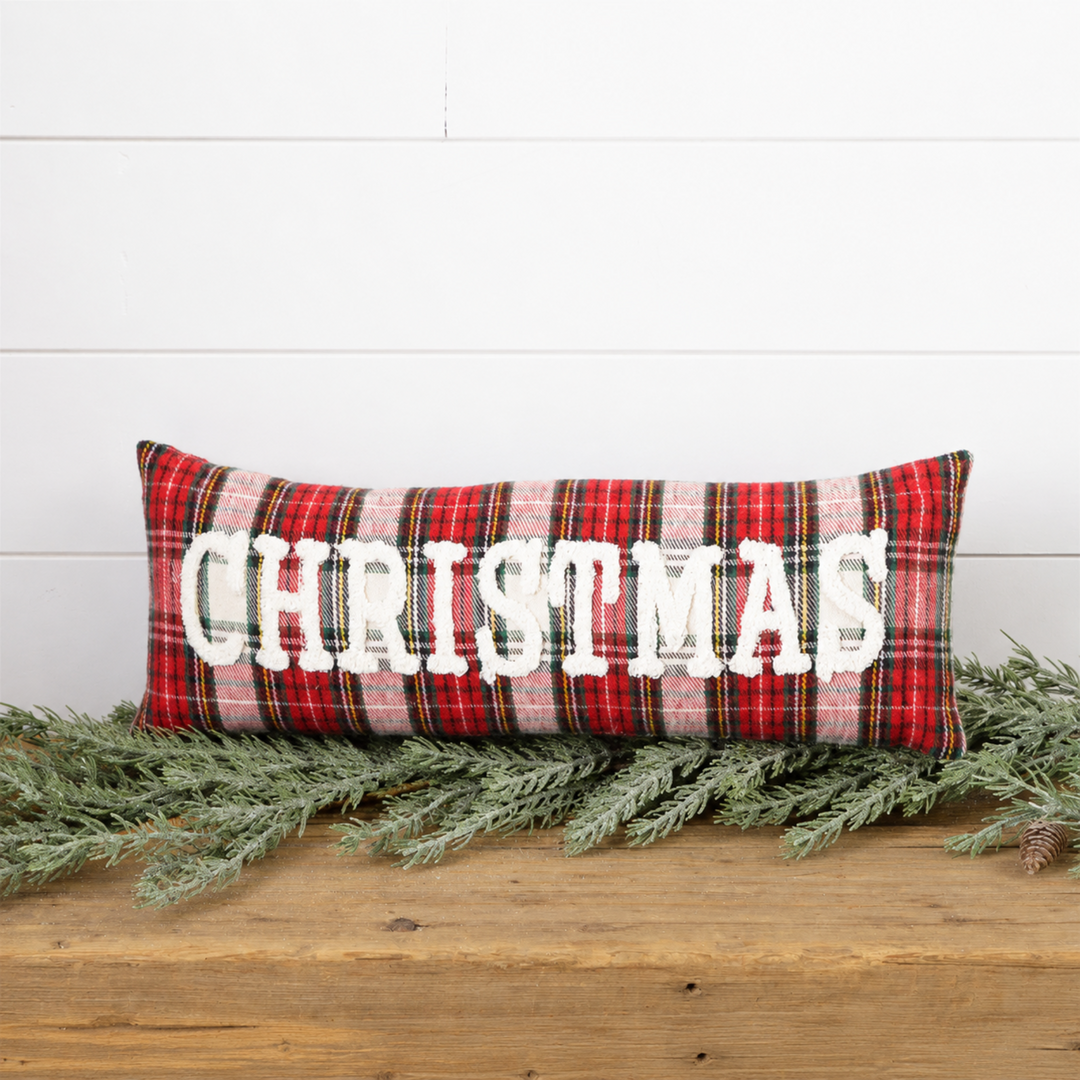 A rectangular holiday throw pillow with a red, green, and white plaid pattern and the word 'CHRISTMAS' in large white letters, styled on a wooden surface with green pine branches against a white shiplap wall.