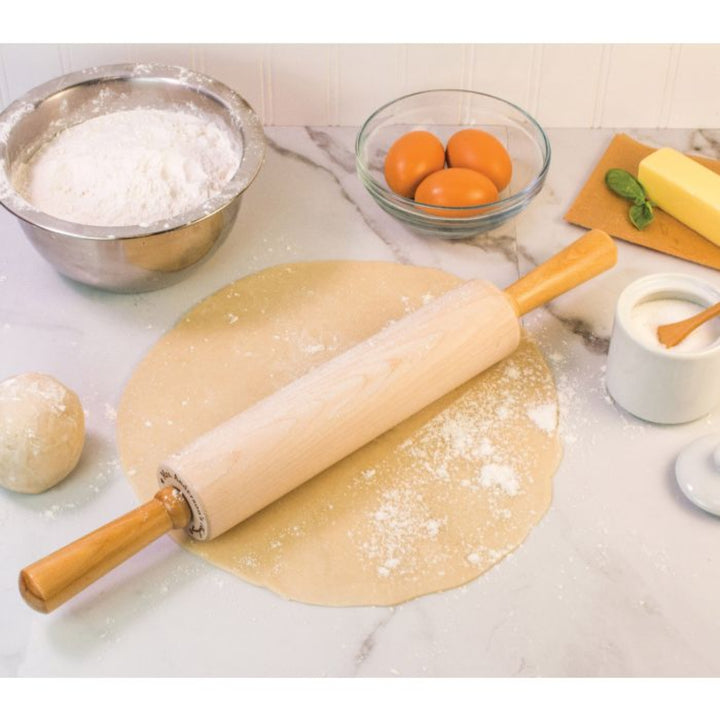 A wooden rolling pin with a silver metal bolster rests on a round of rolled-out dough on a marble countertop, surrounded by baking ingredients including flour, eggs, butter, and salt.