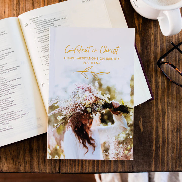 Booklet titled 'Confident in Christ' on a wooden surface with an open book and a cup of coffee.