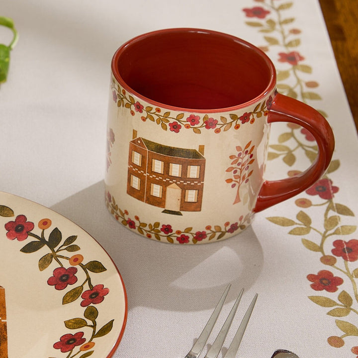 Decorative mug and plate with floral and house design on a matching tablecloth.
