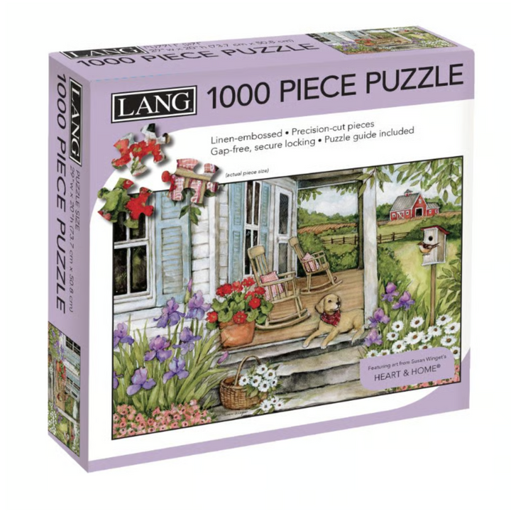 Lang 1000 Piece Puzzle box featuring an illustrated cottage porch scene with flowers, a rocking chair, a dog, and a garden. The box is lavender with black and white text and shows the puzzle name 'Heart & Home'.