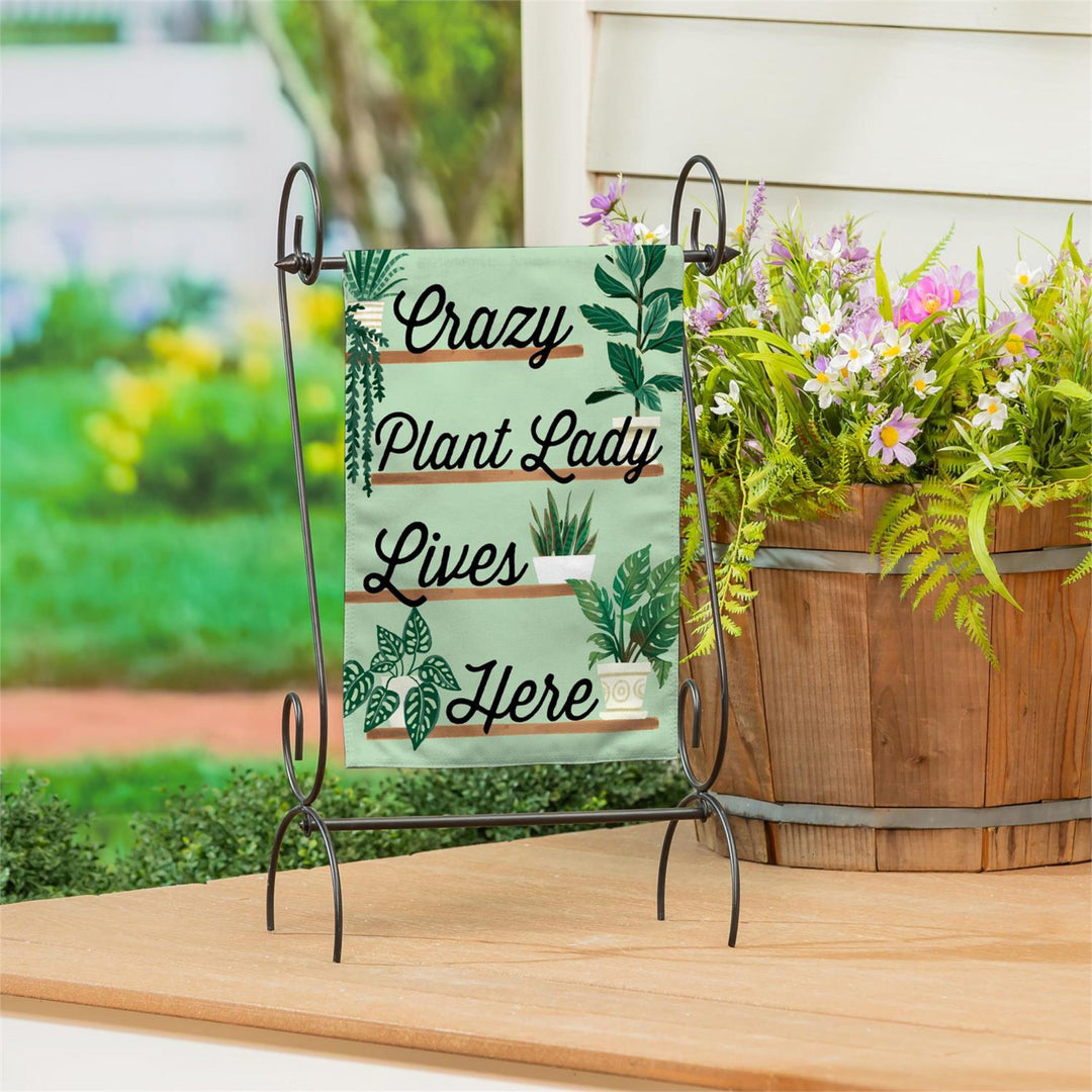 Decorative sign with 'Crazy Plant Lady Lives Here' text on a stand with plants in the background.