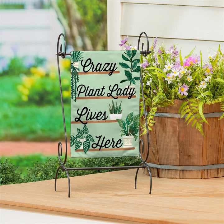 Decorative sign with 'Crazy Plant Lady Lives Here' text on a stand with plants in the background.