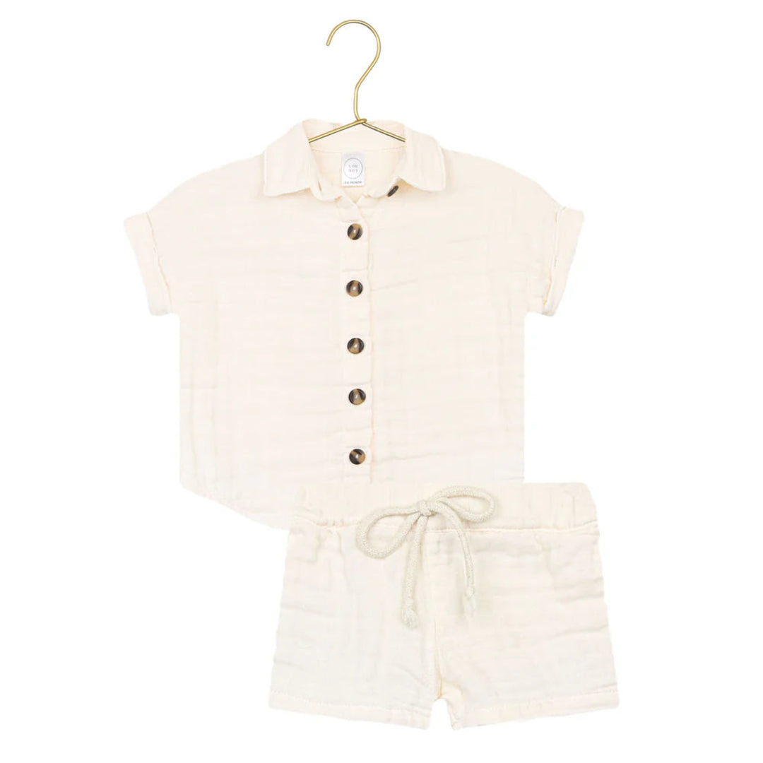 A two-piece baby outfit set in cream-colored muslin fabric, featuring a short-sleeve button-up shirt with a collar and matching drawstring shorts, displayed on a gold hanger against a white background.