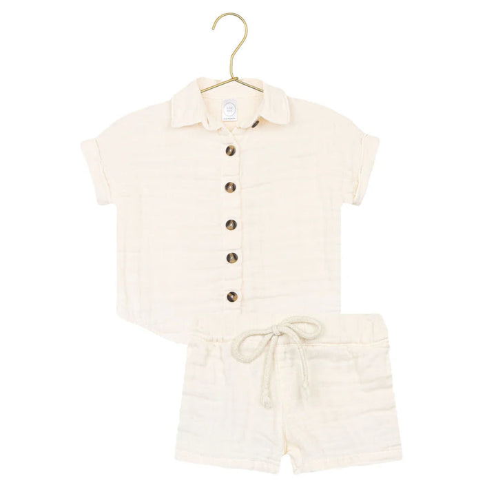 A two-piece baby outfit set in cream-colored muslin fabric, featuring a short-sleeve button-up shirt with a collar and matching drawstring shorts, displayed on a gold hanger against a white background.