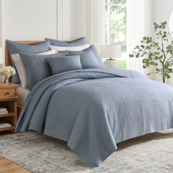 Blue textured bedding set on a bed in a bedroom with a window and plant in the background.