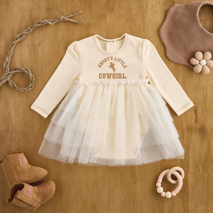 A baby girl's long-sleeve bodysuit dress styled in a flat lay on a brown wooden surface. The dress is cream-colored with 'BERRY'S LITTLE COWGIRL' printed in brown on the chest.