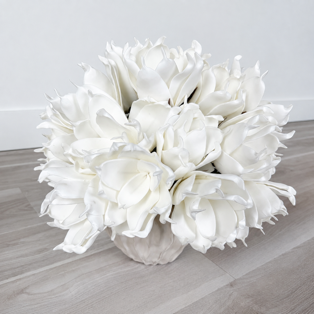 A large bouquet of white artificial flowers arranged in a textured white vase, placed on a light wood floor against a white wall.