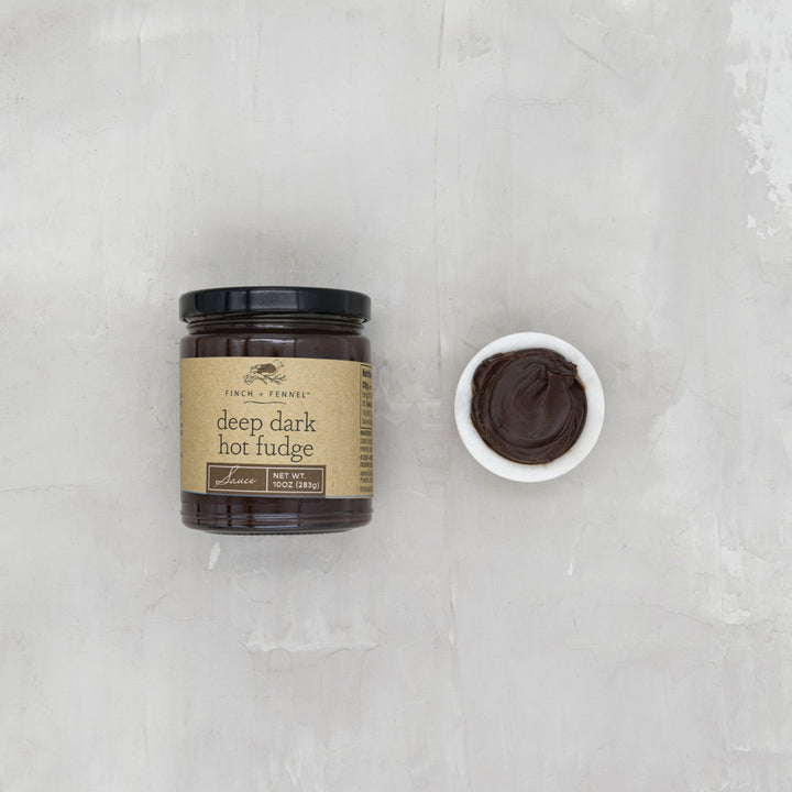 Jar of deep dark hot fudge on a gray surface with a small bowl of fudge beside it.