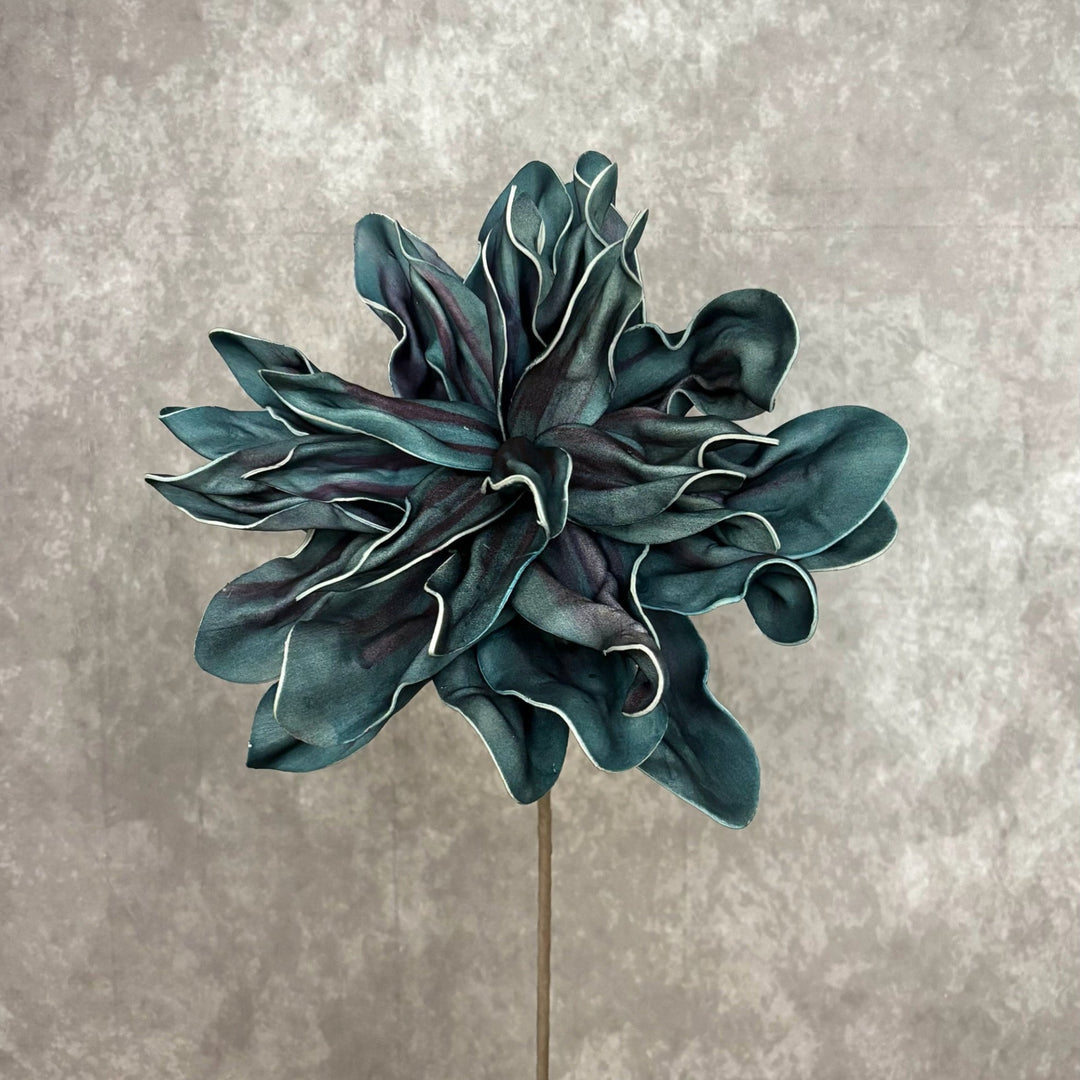 A single sculptural flower head on a thin stem, shown against a textured gray studio background. The petals are a deep teal-blue with darker shadowed areas and thin light edging, mounted on a vertical stem.