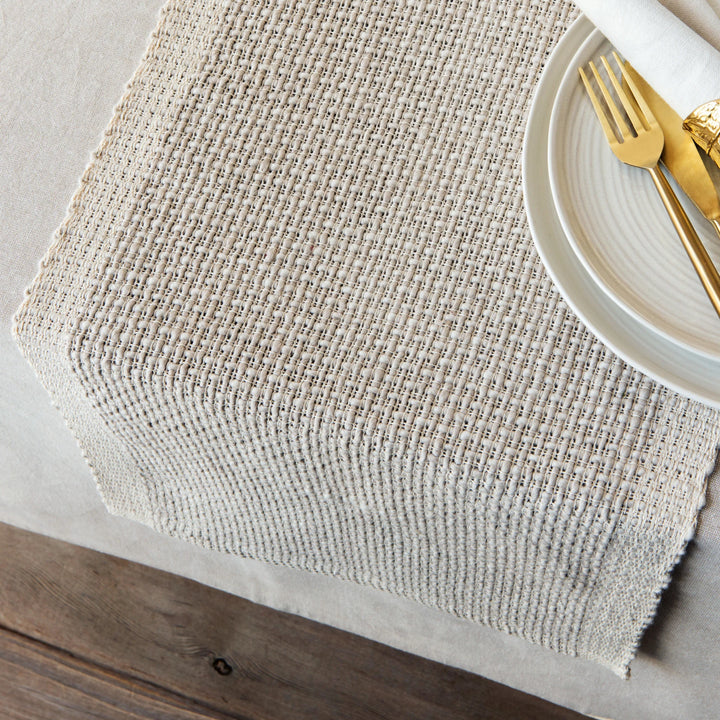 Textured beige placemat on a wooden table with a white plate and gold cutlery.