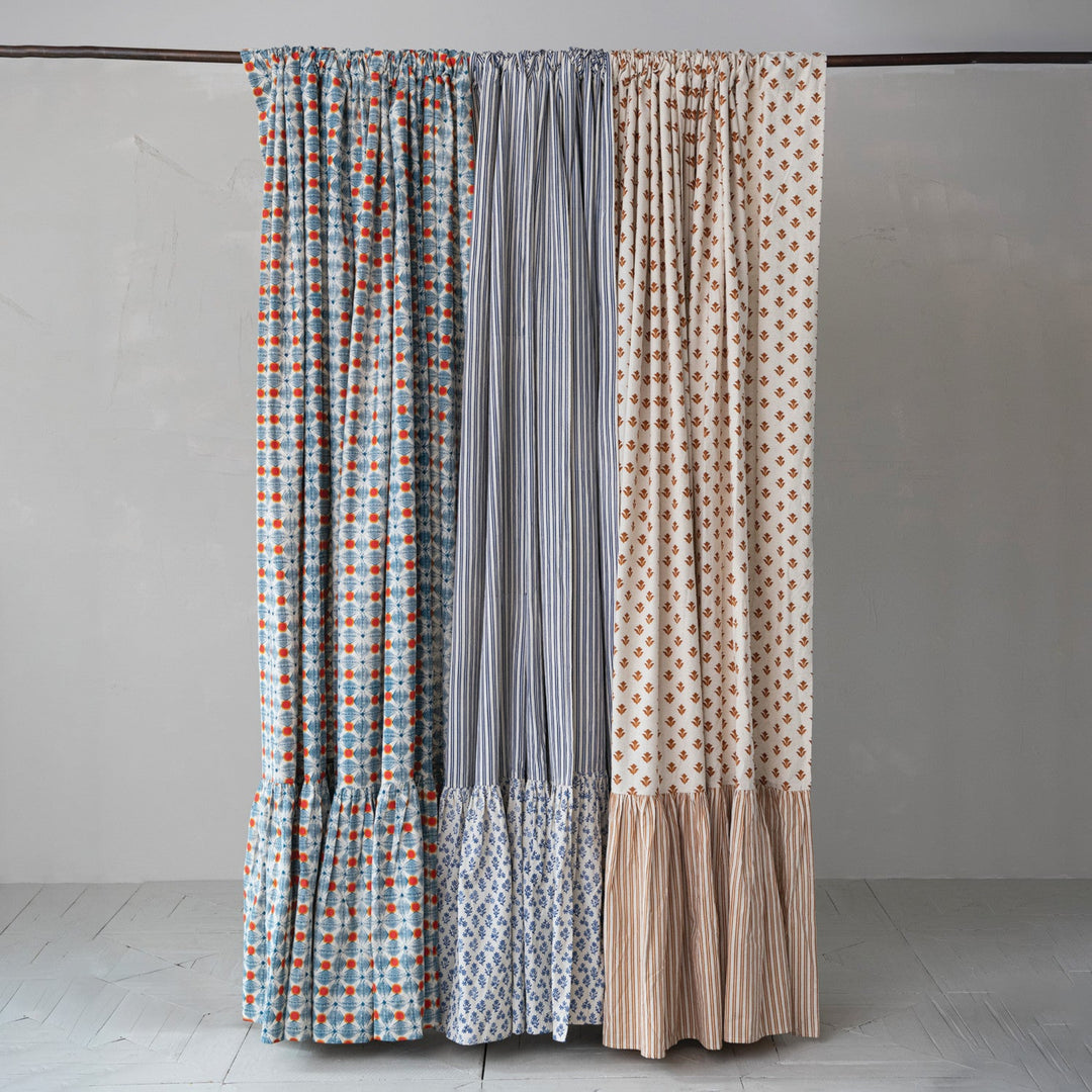 Three patterned curtain panels hanging from a dark rod against a plain light gray wall and pale floor. The left panel has a blue, white, and orange geometric pattern; the center panel is vertically striped in gray and white; the right panel has a beige background with orange geometric dots and a beige striped ruffled hem.